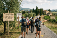 Group of hikers walking on a paved path with backpacks and walking sticks, surrounded by countryside and signage for Camino de Santiago